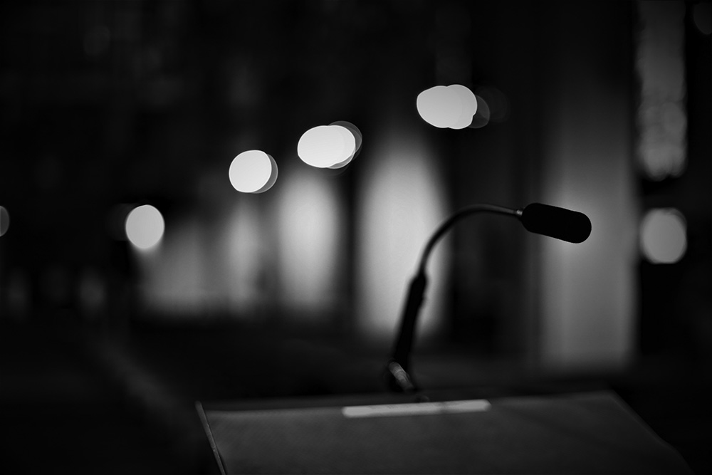Zu sehen ist eine Schwarz-weiß-Fotografie eines Mikrofones auf einem Redner*innenpult. Im Hintergrund sind Wandlichter zu erkennen.