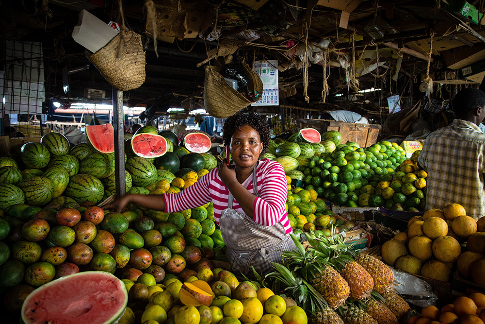 Zu sehen ist eine Frau an einem Marktstand, umgeben von vielen unterschiedlichen Früchten.