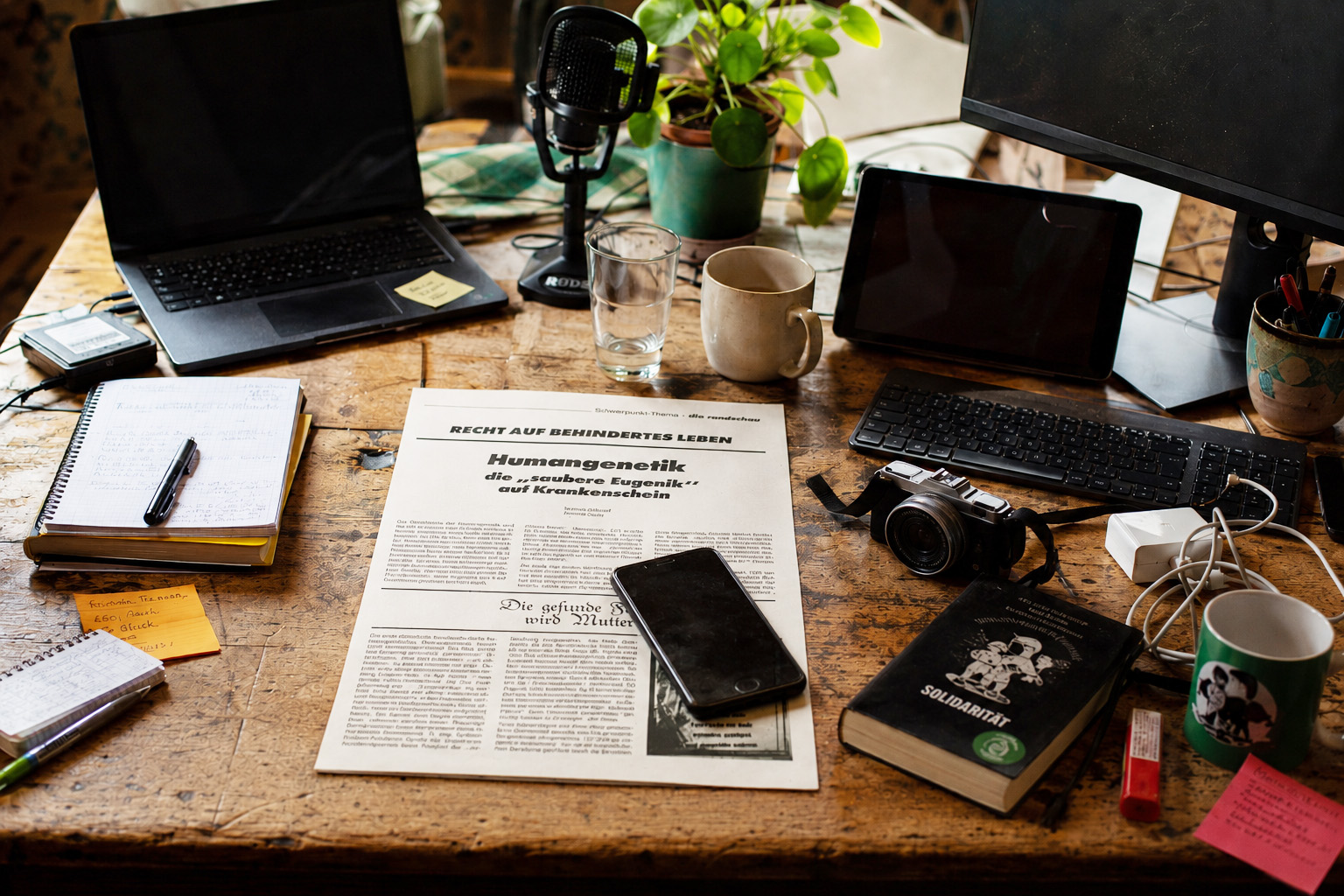Arbeitsplatz mit Laptop, Kamara, Kaffeetasse, Handy und Notizzetteln. Mitten auf dem etwas unübersichtlichen Tisch liegt eine Zeitung mit den Überschriften "Recht auf behindertes Leben" und "Humangenetik - die 'saubere Eugenik' auf Krankenschein"