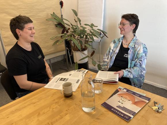 Janina und Laura sitzen an einem Tisch, im Hintergrund steht eine Pflanze, vor ihnen auf dem Tisch liegen zwei GID-Ausgaben und dazwischen steht eine Wasserkaraffe und Gläser