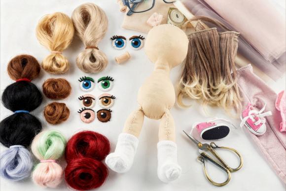 Puppe mit Stoffkörper und weißen Socken umgeben von verschiedenen Haarsträhnen, bunten Puppenaugen, einer Schere, kleinen Schuhen und Nähzubehör auf hellem Untergrund